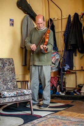 Ein älterer Mann in verblichener Kleidung steht in einem Zimmer seiner Wohnung und spielt auf einer Geige. 